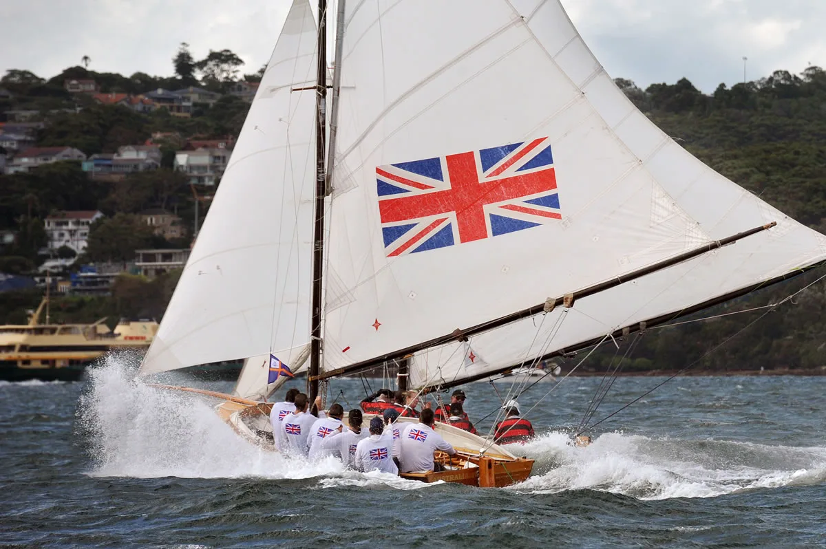 The replica Australia IV heeling through spray on Sydney Harbour, Union Jack insignia on the mainsail and a harbour ferry beyond