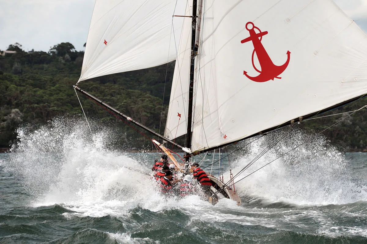 The replica Yendys crashing through spray under full sail, the red anchor insignia on her mainsail and crew in red and black striped jumpers