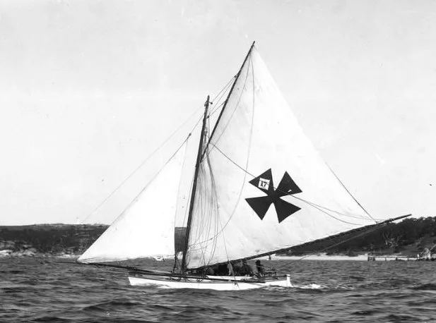 Mark Foy's double hulled Flying Fish sailing under a large gaff mainsail carrying a dark Maltese cross device and the number 17, her crew sitting low in the hull