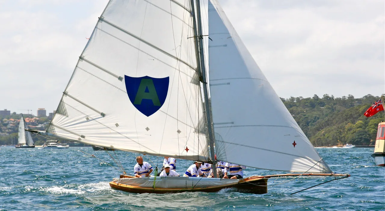 The replica Alruth heeling close-hauled on Sydney Harbour, the blue shield with letter A large on her mainsail and crew in club jerseys along the gunwale