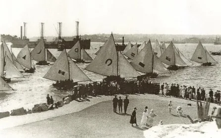 A fleet of open boats carrying large dark devices on their mainsails, photographed from a harbour foreshore where a crowd of men and women in hats stands watching, with steamers moored beyond