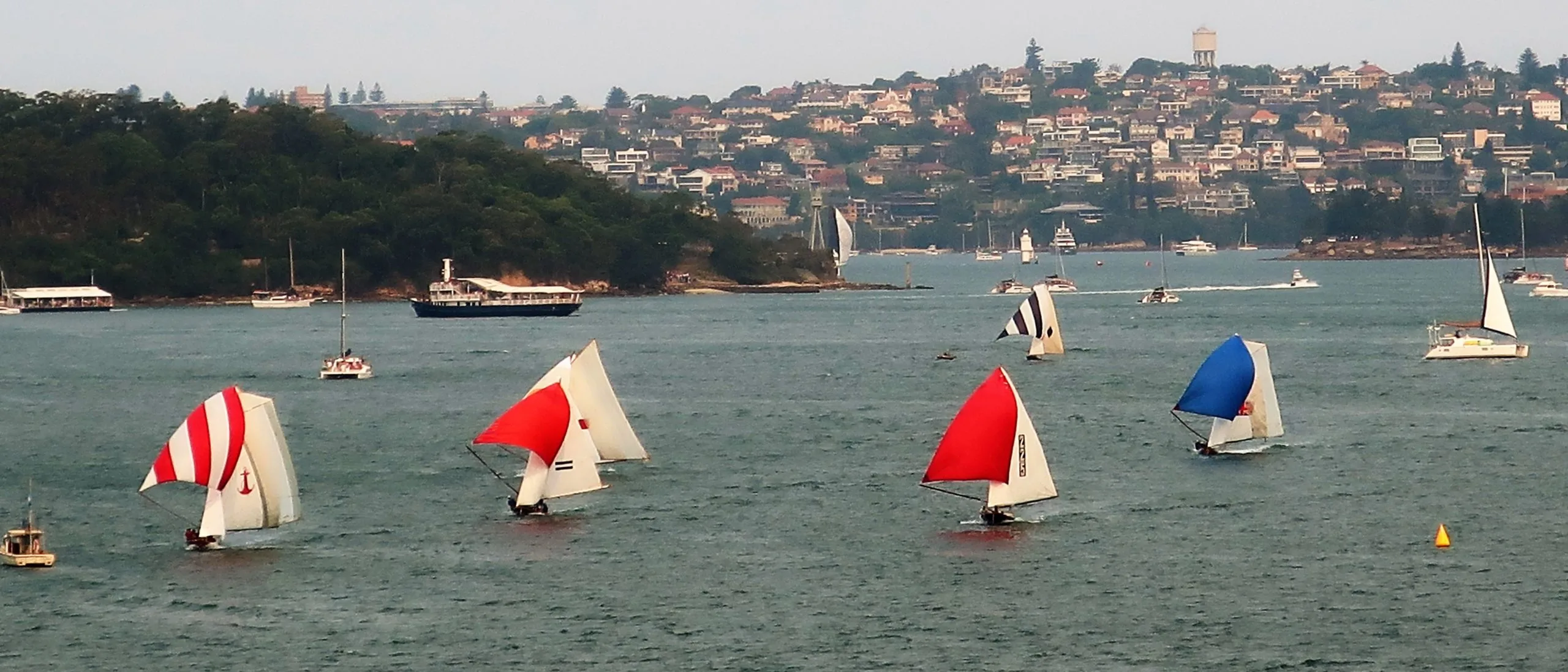 Historic 18-footers running under red-striped, solid red and blue ringtails past the spectator launch off Watsons Bay